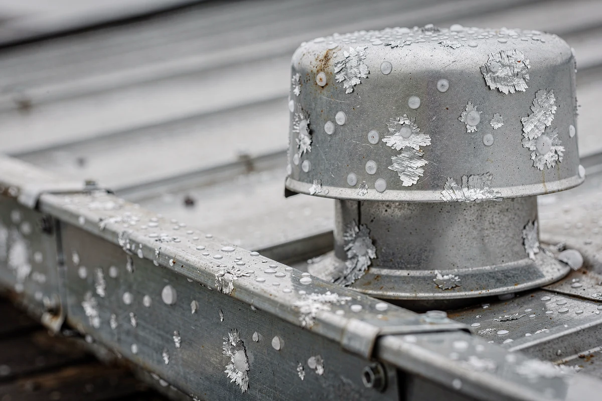 A galvanised roof vent cap and the metal edge below it, both pitted with small round impact marks and flaking coating.