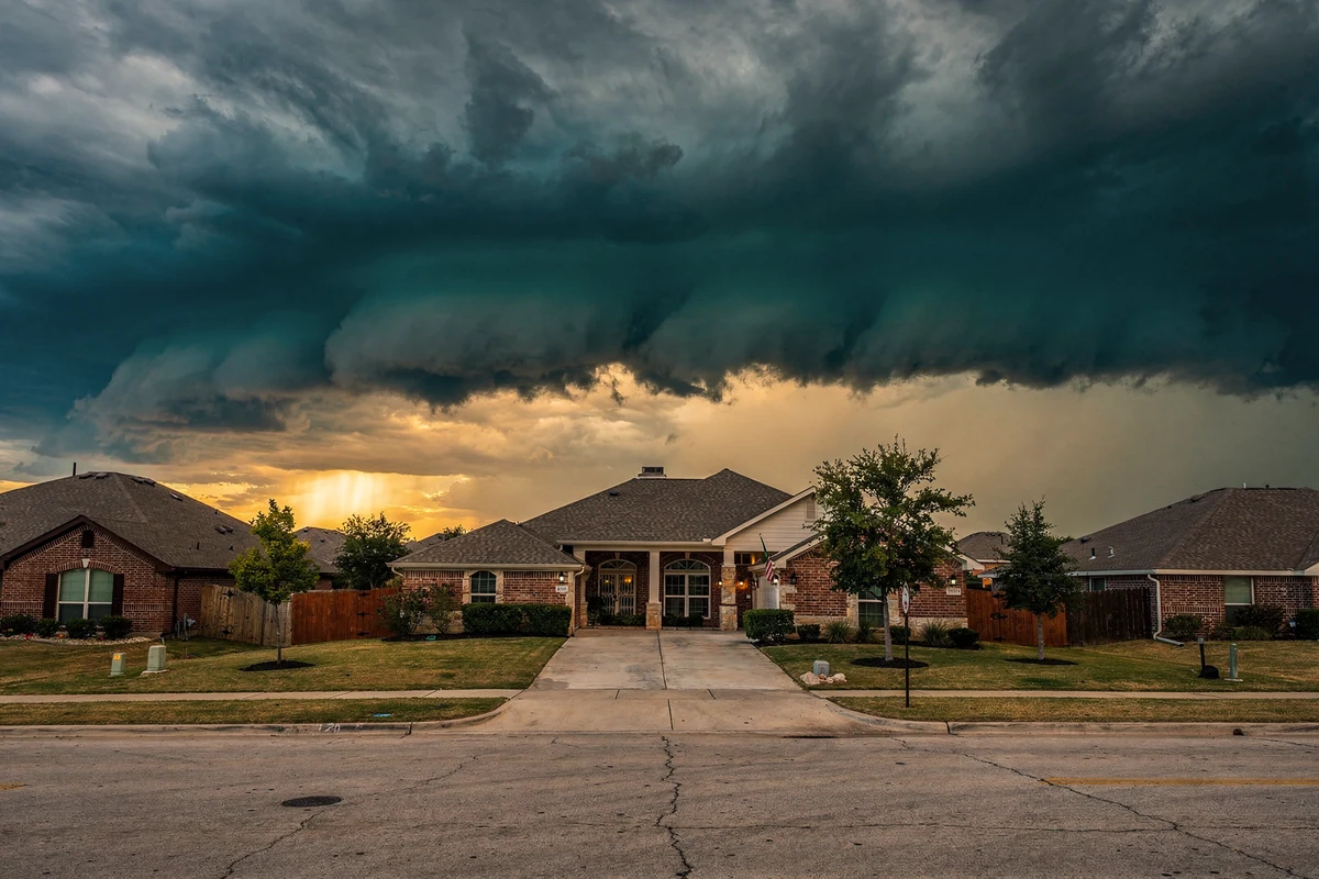 A dark shelf cloud spread across the sky above a straight subdivision street of brick houses with shingle roofs.